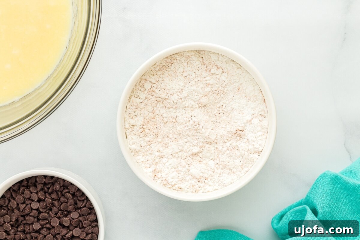 A bowl of gluten-free flour next to a bowl of mini chocolate chips and a glass bowl containing whisked wet muffin ingredients, all arranged on a white surface with a teal kitchen towel.