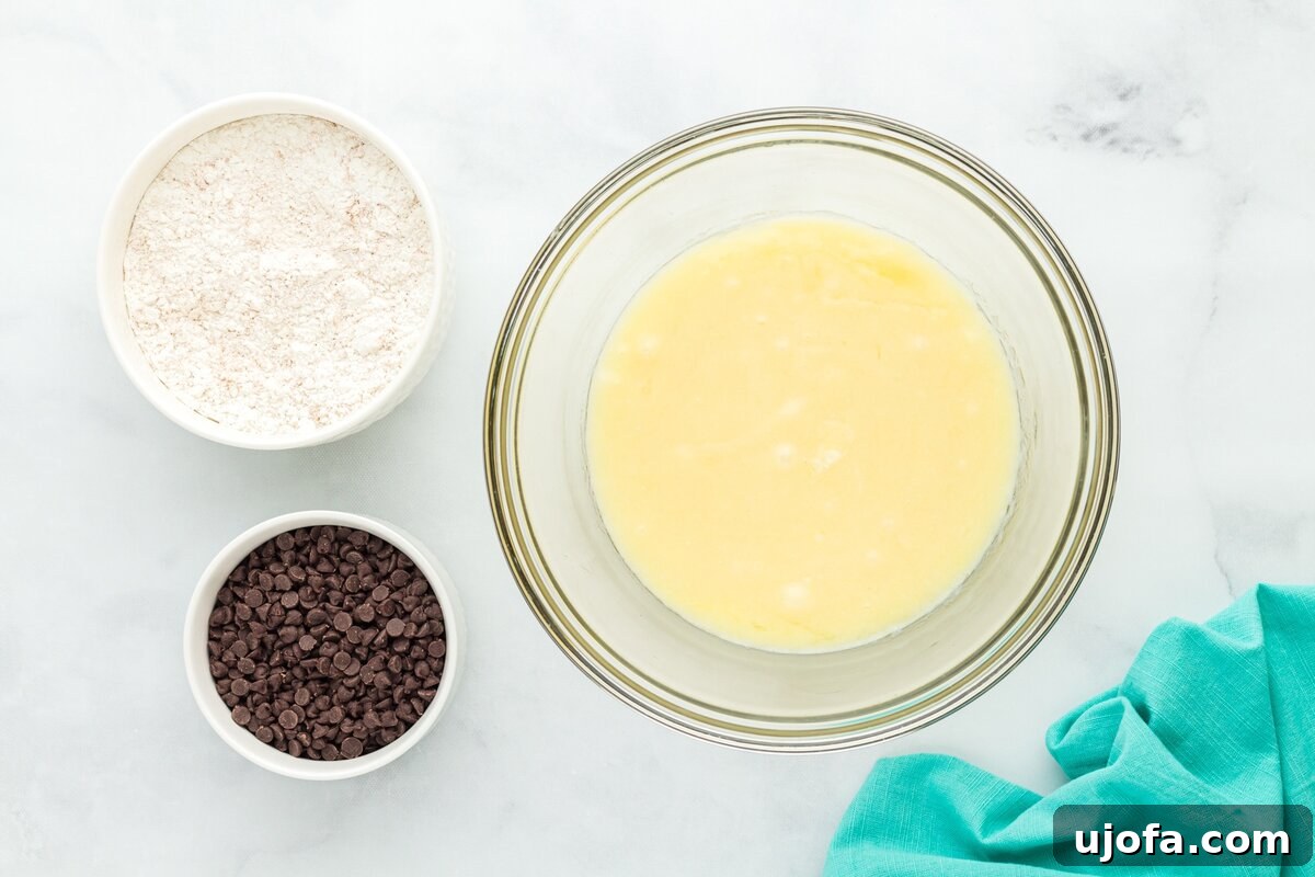 An overhead view showcasing the separated muffin ingredients: a bowl of gluten-free flour, a bowl of mini chocolate chips, and a larger glass bowl holding the mixed wet ingredients (sugar, sour cream, eggs, melted butter, oil, vanilla).