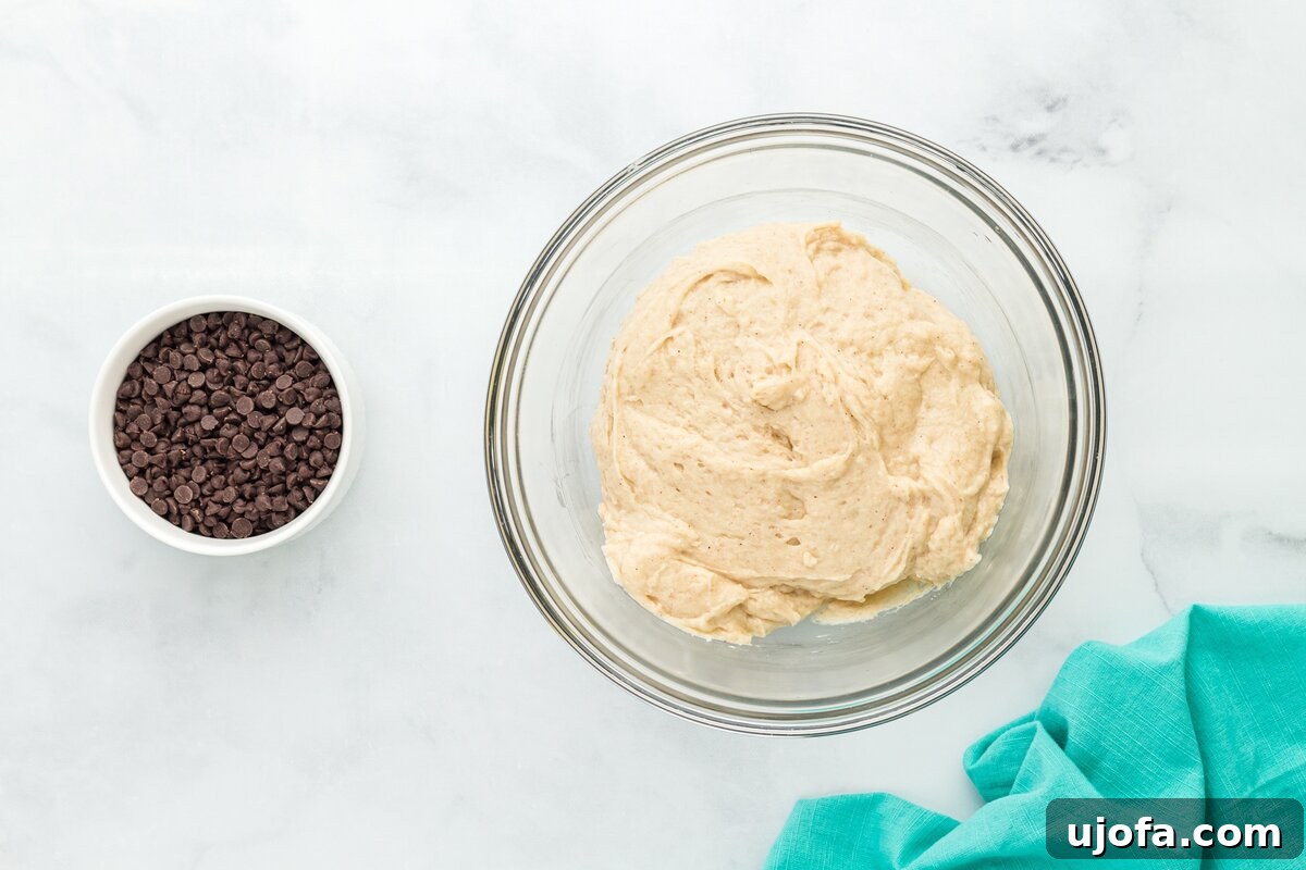A bowl of partially mixed muffin batter, with some dry streaks still visible, sitting next to a separate bowl of mini chocolate chips, waiting to be combined.