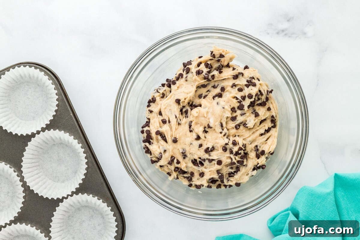 A bowl of fully mixed muffin batter, generously studded with mini chocolate chips, positioned beside a muffin tin already lined with paper wrappers, ready for filling.