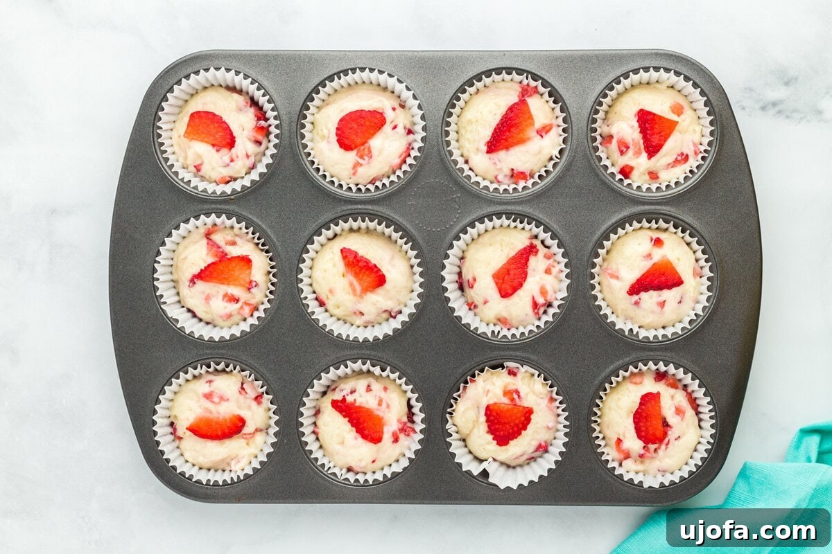 A muffin tin with all cups filled with strawberry muffin batter, some topped with thin slices of strawberry for decoration, ready to rest before baking.