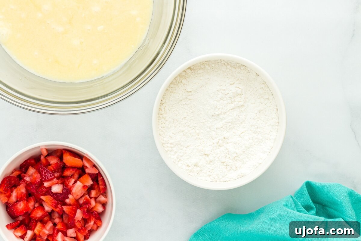 Three bowls on a light background: one with chopped fresh strawberries, one with gluten-free flour mixture, and one with a whisked wet mixture ready for combining.