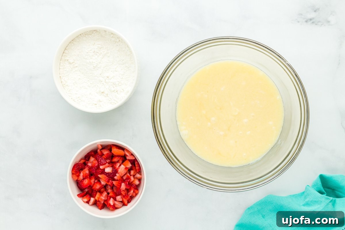 Top-down view of three bowls containing chopped strawberries, gluten-free flour, and a smooth, combined wet muffin batter, set on a white surface.