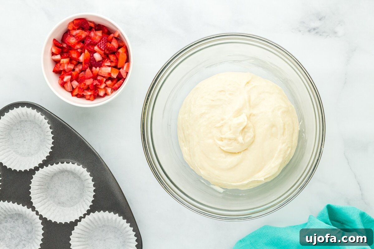 A bowl of thick muffin batter next to a bowl of finely chopped strawberries and a lined muffin pan, all components ready for assembly.
