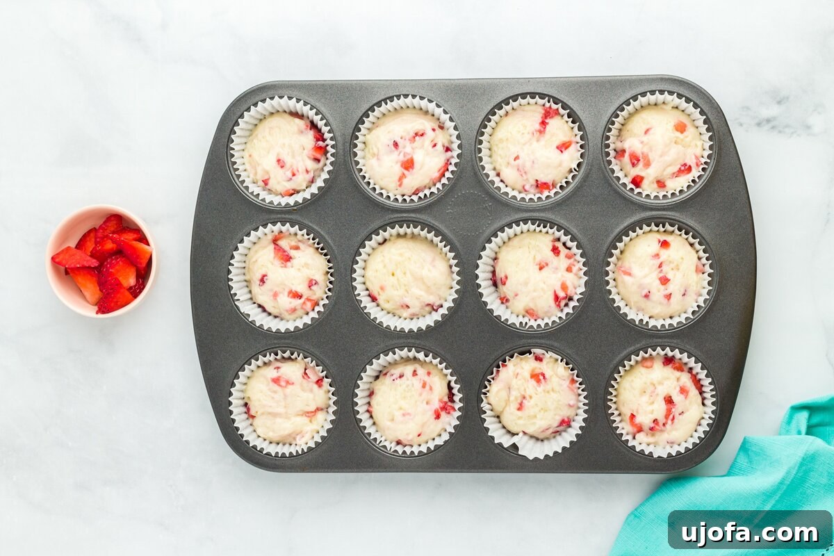A 12-cup muffin pan filled with strawberry muffin batter, each cup approximately three-quarters full and lined with paper baking cups, ready for the oven.