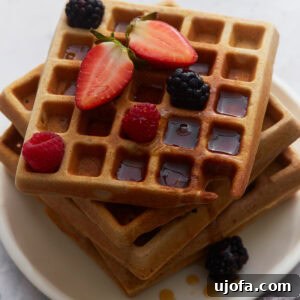 Stacked buckwheat waffles topped with strawberries, raspberries, blackberries, and maple syrup on a white plate, ready for a delicious breakfast.