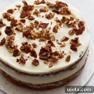 Almond flour carrot cake layered and frosted with cream cheese frosting, topped with chopped pecans on a white cake stand.