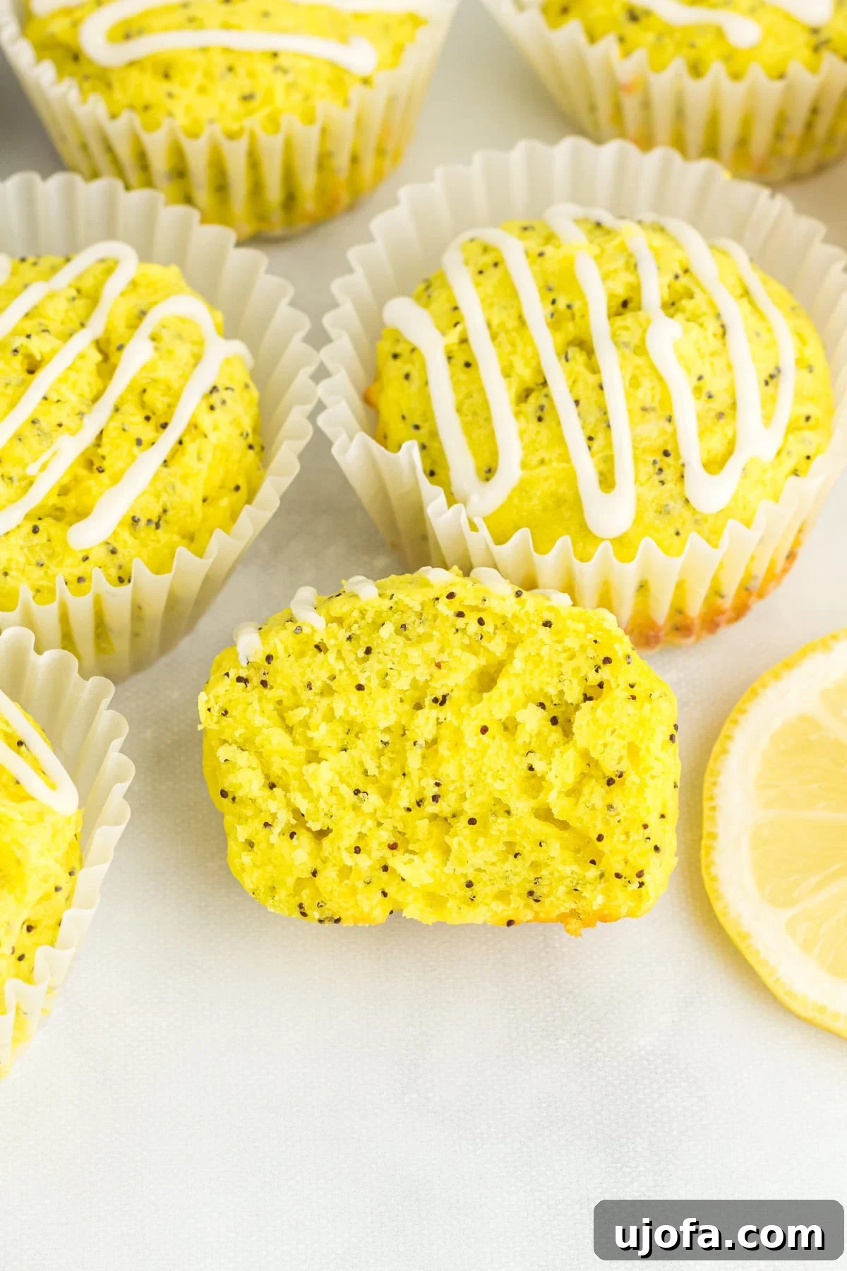 Gluten-free lemon poppy seed muffins with lemon glaze in white liners, one muffin sliced open to show moist crumb and poppy seeds