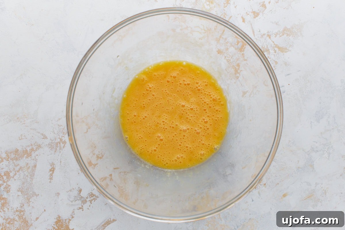 Artisanal Almond Biscotti 4 Two large eggs and melted unsalted butter are whisked together in a clear glass mixing bowl, creating a smooth, yellow liquid mixture.