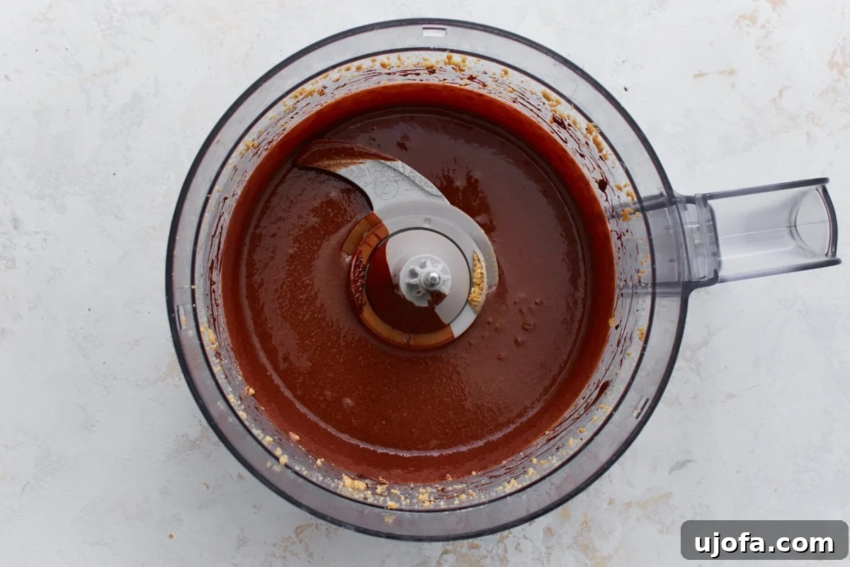 A close-up of the chocolate hazelnut mixture in a food processor, appearing smooth and glossy after the addition of melted dark vegan chocolate, vanilla extract, and salt.