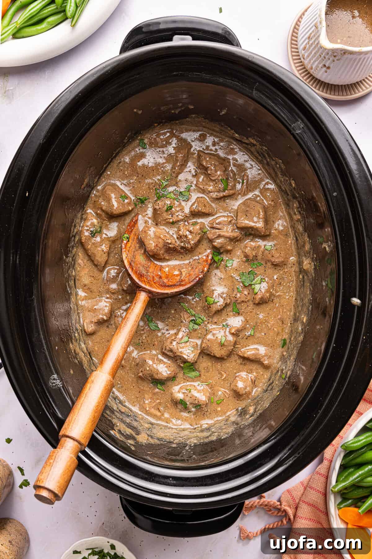 slow cooker beef tips with gravy topped with parsley in a crock pot with a wooden spoon