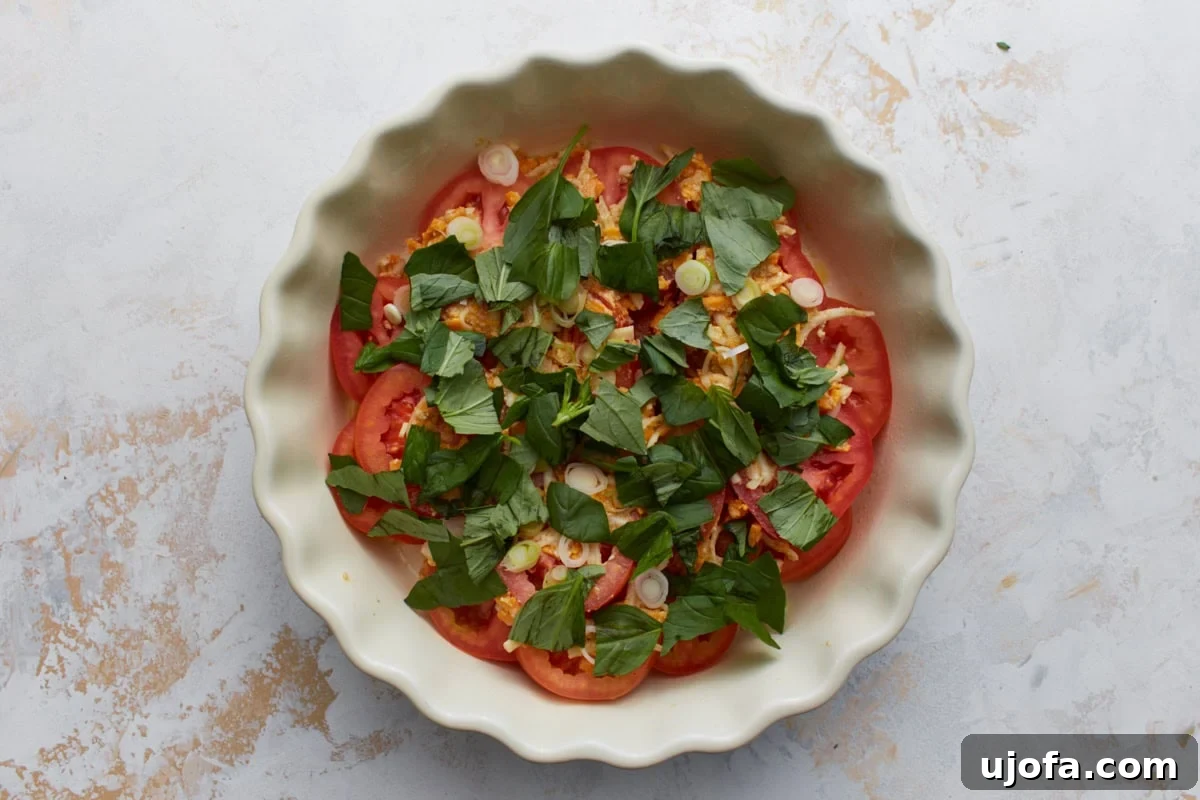 Overhead view of crustless tomato pie before baking with layers of sliced tomatoes, cheese mixture, fresh basil, and green onions in a white pie dish.