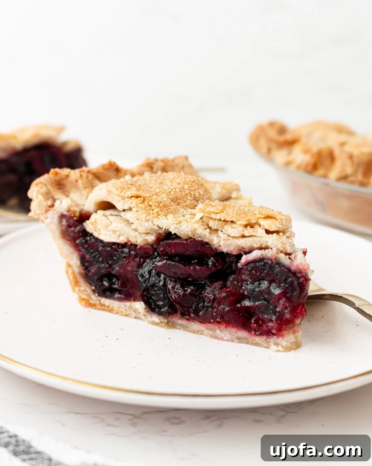 A close-up side view of a slice of gluten-free cherry pie on a white plate with a gold rim, showing a flaky golden lattice crust and a deep, jammy cherry filling, with the whole pie visible in the background.