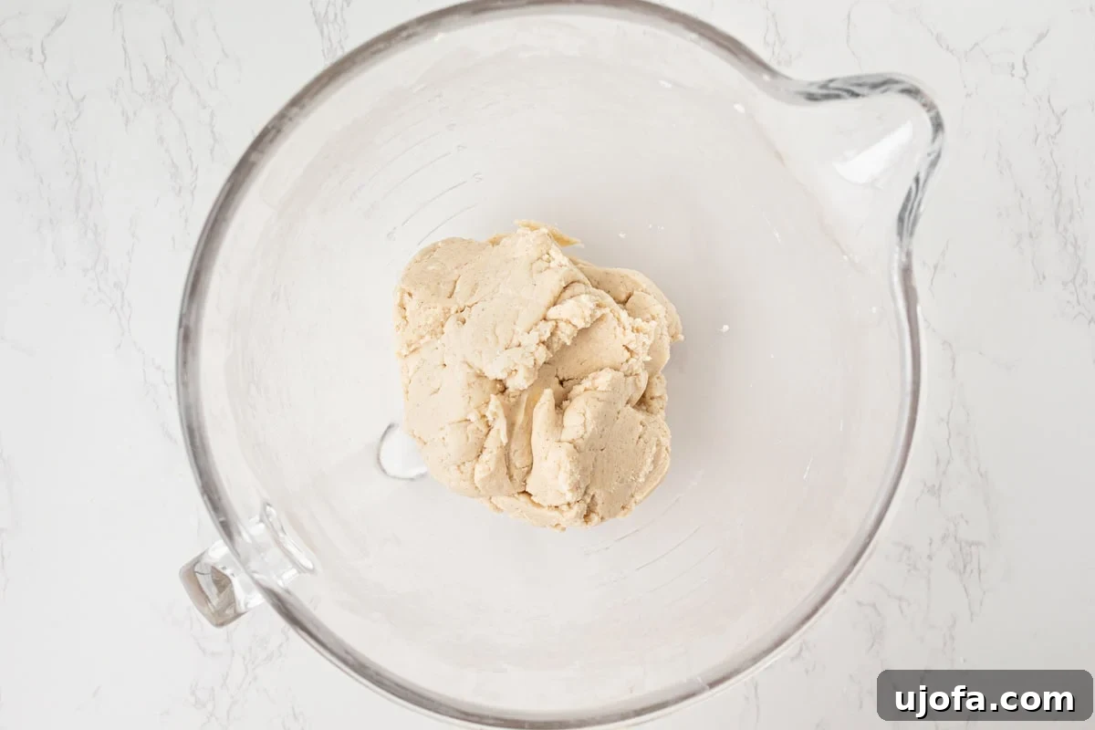 Overhead view of gluten-free pie crust dough formed into a rough ball in a glass stand mixer bowl on a white marble surface.