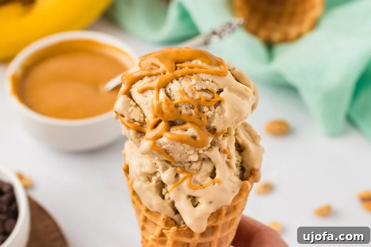 Hand holding a waffle cone with two scoops of banana peanut butter nice cream drizzled with melted peanut butter