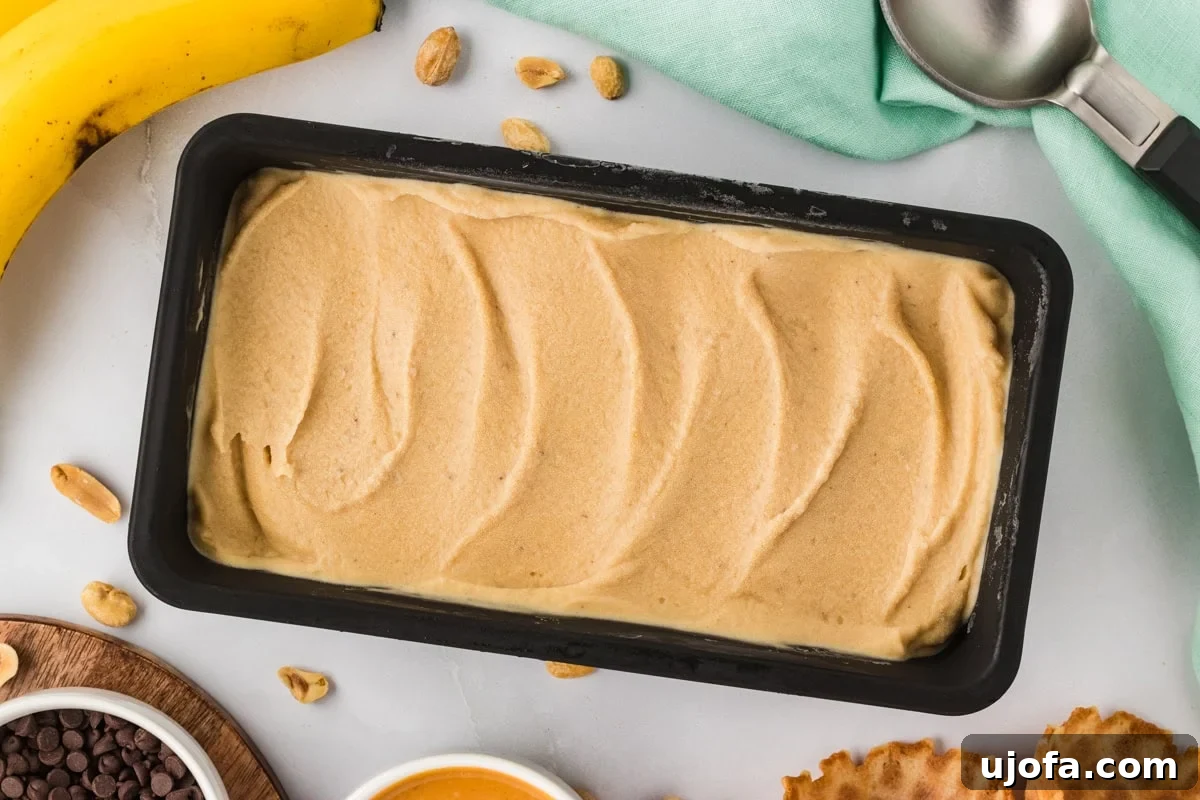 Overhead view of banana peanut butter nice cream in a black loaf pan surrounded by peanuts, chocolate chips, a waffle cone, and a banana