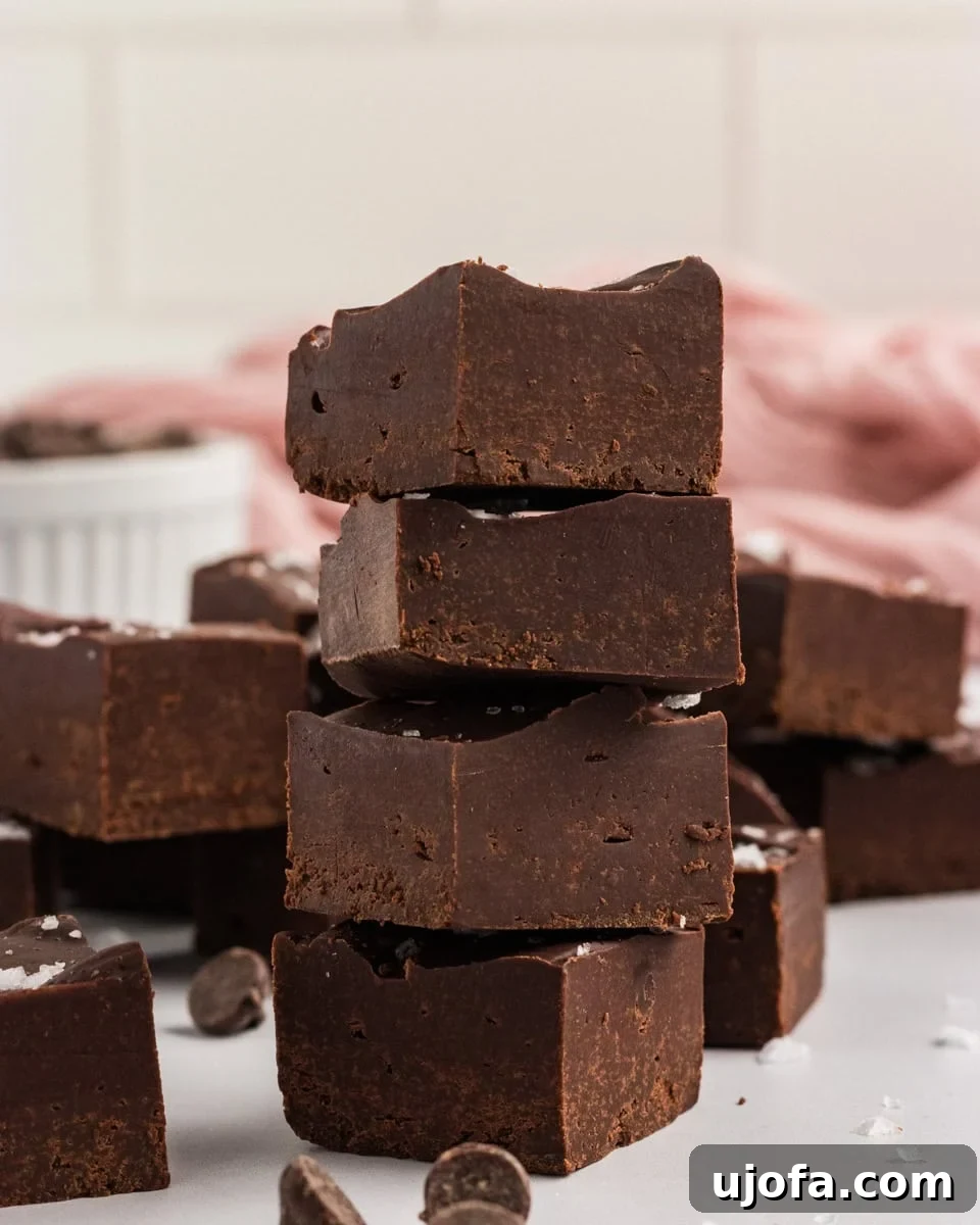 Rich Gluten-Free Dairy-Free Chocolate Fudge Recipe 2 Stack of gluten-free dairy-free chocolate fudge squares topped with flaky sea salt, with chocolate chips scattered around and a pink cloth in the background