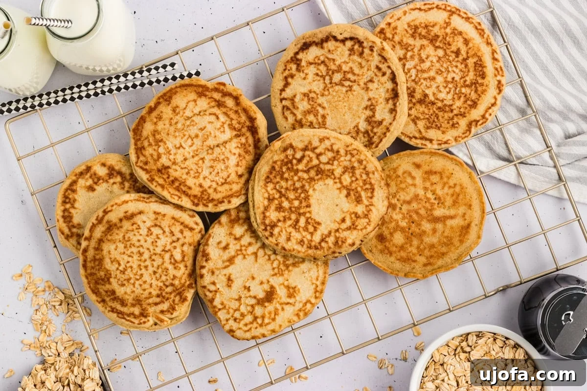 Fluffy Oat Flour Pancakes Recipe 9 Cooked oat flour pancakes cooling on a wire rack next to milk bottles and a bowl of rolled oats.