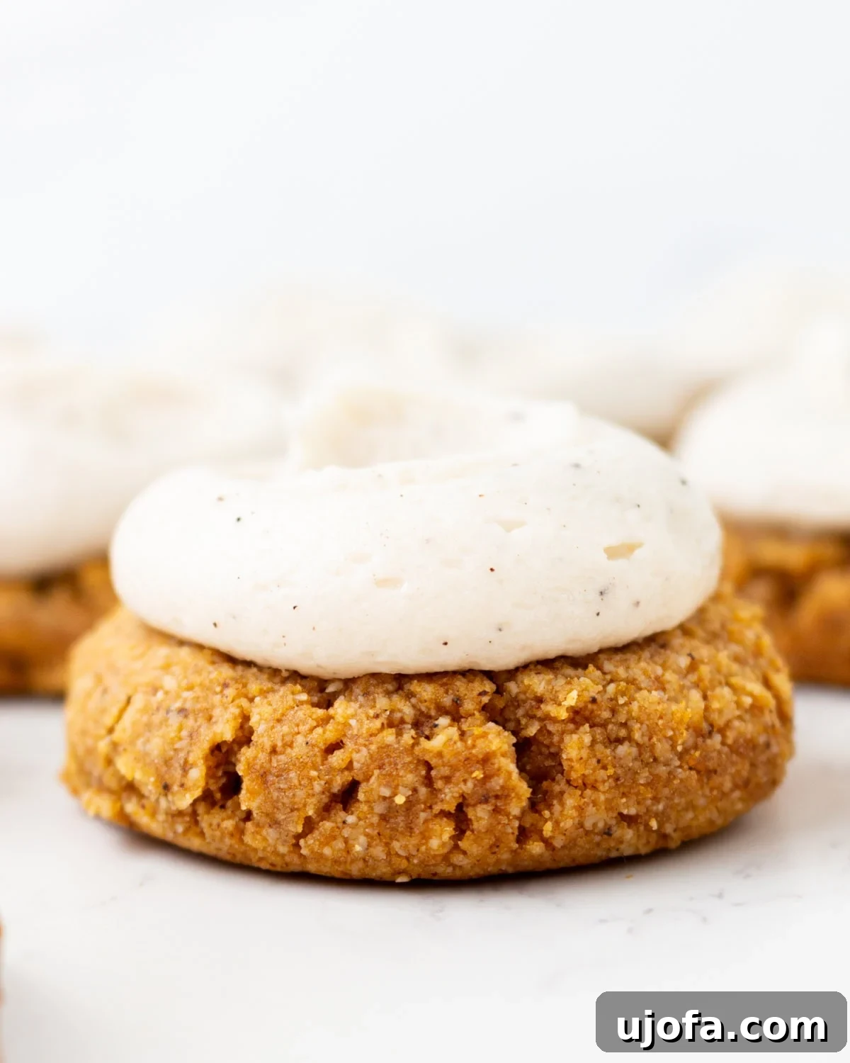 Chewy Almond Flour Pumpkin Cookies with Warm Spices 2 Gluten-free almond flour pumpkin cookie topped with brown butter cream cheese frosting, with more frosted cookies blurred in the background.